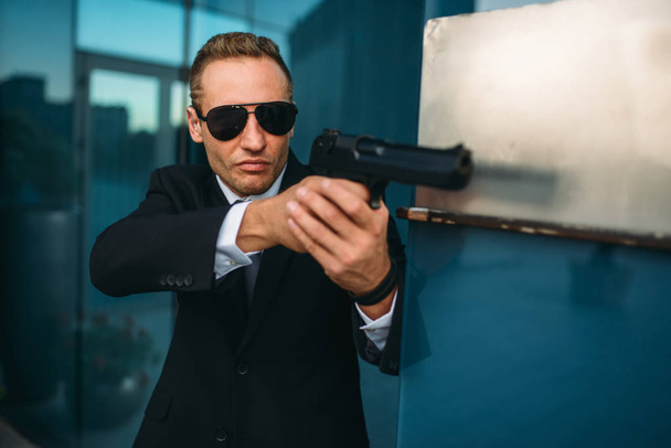 Bodyguard in suit and sunglasses with a gun in his hands. Security guard is a risky profession, professional guarding, politicians and business persons protection from the danger of life - Photo, Image