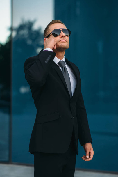 Male bodyguard talking by earpiece outdoors, security communication tools. Professional guarding is a risky profession. Guard in suit and sunglasses - Photo, Image