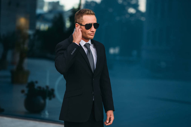 Male bodyguard uses security earpiece outdoors, professional communication tools. Guarding is a risky profession. Guard in suit and sunglasses - Photo, Image