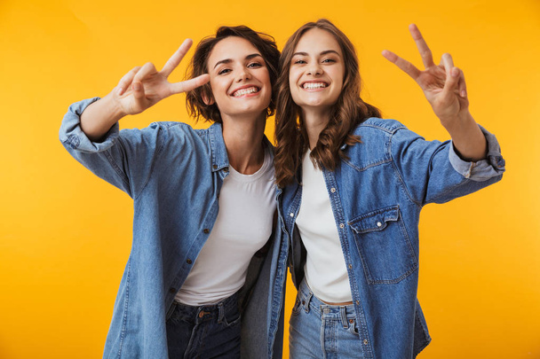 Image of happy emotional young women friends posing isolated over yellow background showing peace gesture. - Foto, immagini