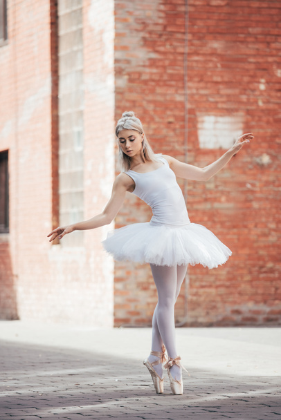 attractive young ballerina in white tutu and pointe shoes dancing on street - Foto, imagen