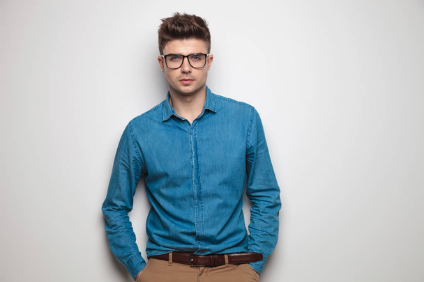 portrait of relaxed casual man wearing sunglasses and blue shirt while standing near a light grey wall with hands in pockets - Foto, Bild