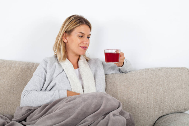 Beautiful woman relaxing on the sofa and drinking hot tea, wrapped in a grey blanket - Foto, Bild