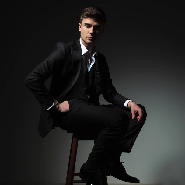 side view of elegant man in black tuxedo resting on wooden chair on grey background with a hand in pocket - Foto, imagen