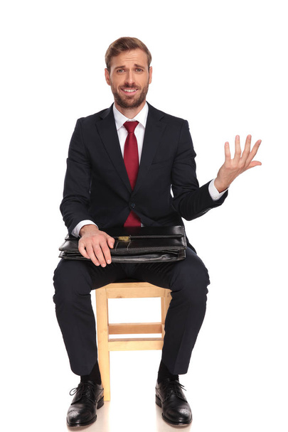 саркастичный бизнесмен ждет интервью, делая жест открытой руки, сидя на белом фоне, полная картина тела
 - Фото, изображение