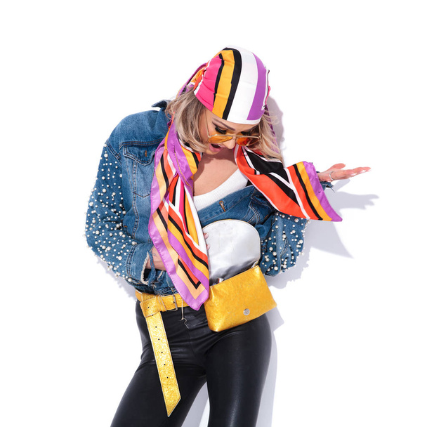portrait of surprised young woman wearing colorful clothes looking down in her waist bag while standing near a white wall - Photo, Image