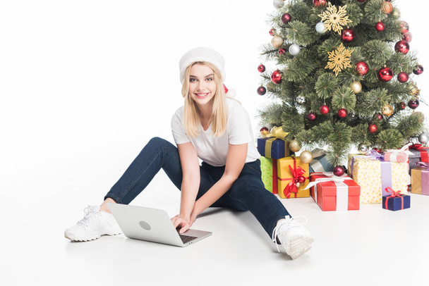 smiling woman in santa claus hat with laptop sitting near christmas tree on white background - Zdjęcie, obraz