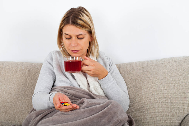 Young woman having seasonal influenza is resting on thesofa, drinking hot tea and taking medication - Фото, изображение