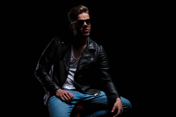 man wearing leather jacket and sunglasses sits on wooden chair and looks up to side on black background, portrait picture - Photo, Image