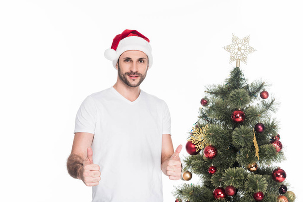 portrait of young man in santa claus hat showing thumbs up near christmas tree isolated on white - Valokuva, kuva