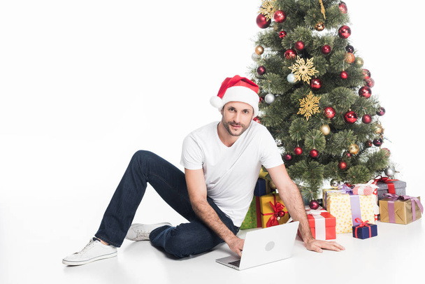 man in santa claus hat with laptop sitting near christmas tree isolated on white - Фото, зображення