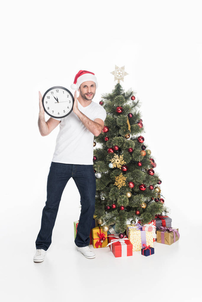 man in santa claus hat with clock standing near christmas tree isolated on white - Foto, Imagen