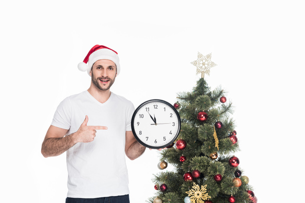 Portret mężczyzny w czapki Mikołajów, wskazując na zegar, podczas gdy na białym tle stojącego obok choinki - Zdjęcie, obraz