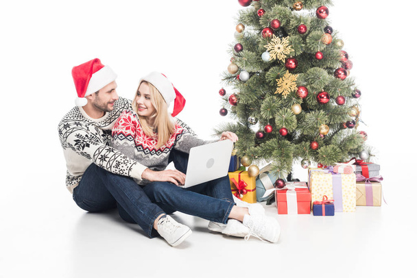 smiling couple in santa claus hats with laptop near christmas tree isolated on white - Foto, Imagen