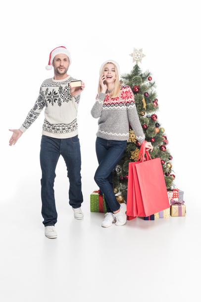 couple in sweaters and santa claus hats with shopping bags near christmas tree isolated on white - Foto, Bild
