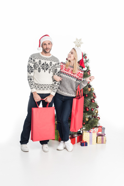 couple in sweaters and santa claus hats with shopping bags near christmas tree isolated on white - Foto, Imagem