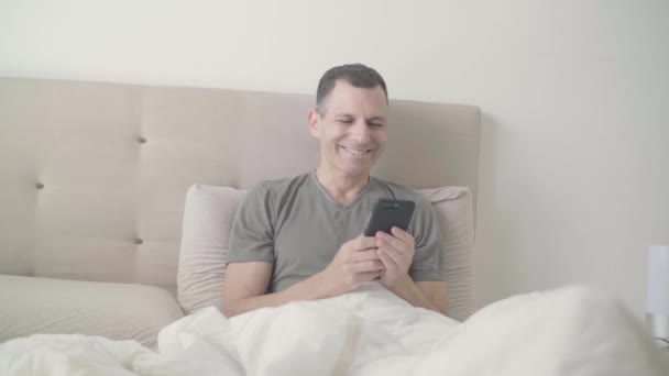 Happy man in bed texting on his smartphone - Materiał filmowy, wideo