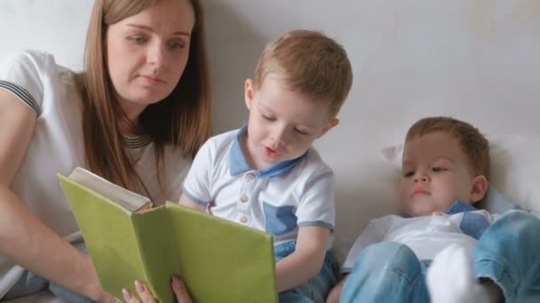 Family mom and two twin brothers toddlers read books laying on the bed. Family reading time. - Video