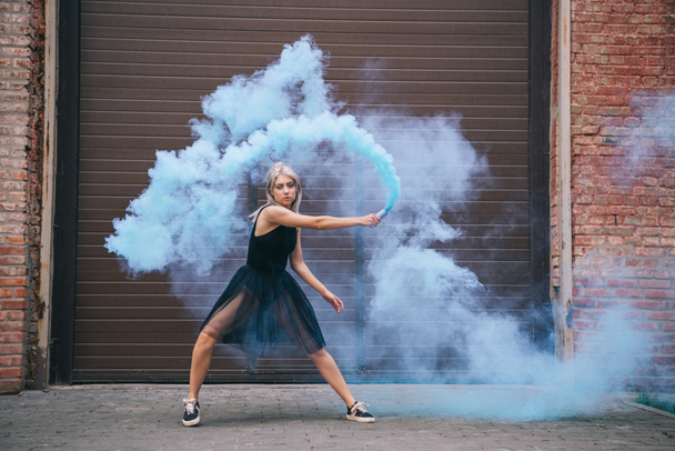 attractive girl looking at camera and dancing in blue smoke on street - Foto, imagen