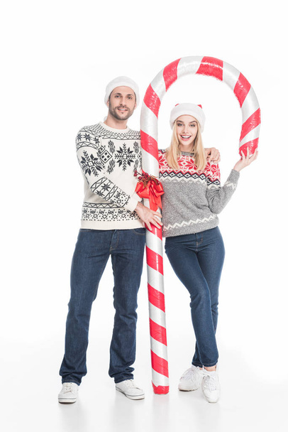 smiling couple in santa claus hats and sweaters with toy candy isolated on white - 写真・画像