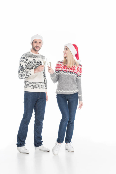 young couple in sweaters and santa claus hats clinking glasses of champagne isolated on white - Foto, Imagen