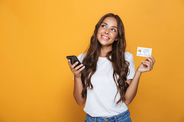 黄色の背景の上に立って、携帯電話を保持している、プラスチック製のクレジット カードを示す長いブルネットの髪を持つ美しい若い女の子の肖像画 - 写真・画像