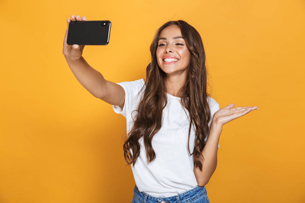 黄色の背景に分離されたスマート フォンで selfie 写真を取っている間長い髪が笑っていると肯定的な女性 20 代のポートレート - 写真・画像