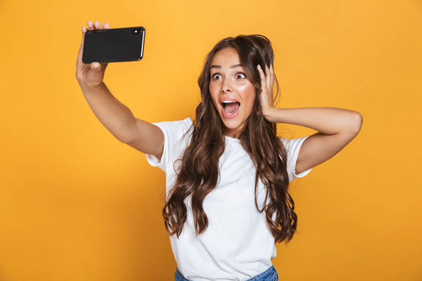 Portrait of caucasian woman 20s with long hair yelling in surprise while taking selfie photo on smartphone isolated over yellow background - Fotoğraf, Görsel