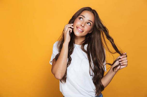 Portrait of gorgeous woman 20s with long hair smiling and talking on cell phone isolated over yellow background - Фото, изображение