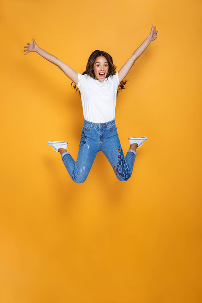Full length portrait of a cheerful girl with long dark hair jumping over yellow background, celebrating success - Φωτογραφία, εικόνα