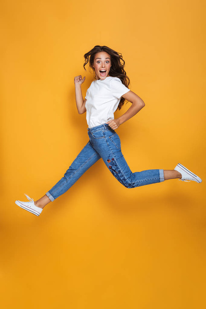Full length portrait of a cheerful girl with long dark hair jumping over yellow background, looking at camera - Foto, imagen