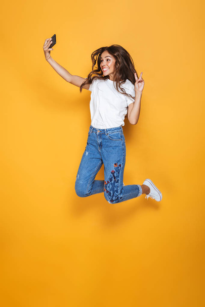 Full length portrait of a happy girl with long dark hair jumping over yellow background, taking a selfie - Foto, Bild