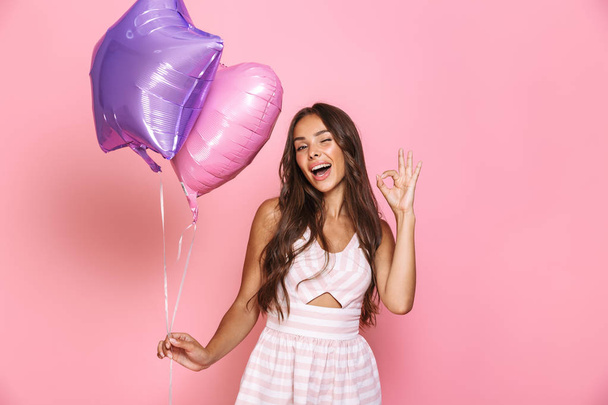 Portrait of charming young woman 20s with long hair wearing dress smiling and holding two balloons isolated over pink background - Foto, Imagen