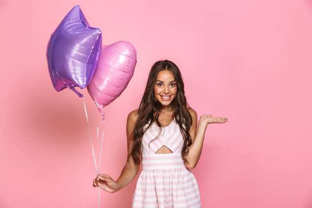 Portrait of charming brunette girl 20s with long hair wearing dress smiling and holding two balloons isolated over pink background - Zdjęcie, obraz