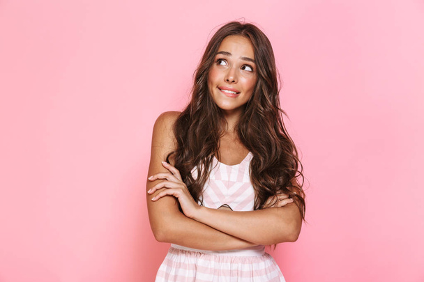 Image of charming woman 20s with long hair wearing dress smiling aside with arms crossed isolated over pink background - Photo, Image