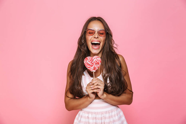Photo of fashion woman 20s wearing sunglasses laughing and eating lollipop isolated over pink background - Foto, Bild