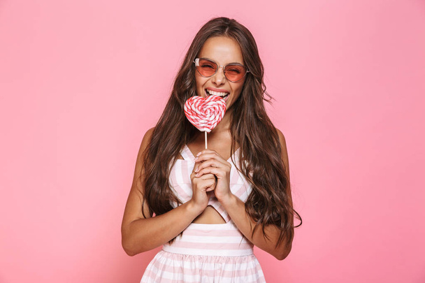 Photo of young woman 20s wearing sunglasses laughing and eating lollipop isolated over pink background - Photo, Image