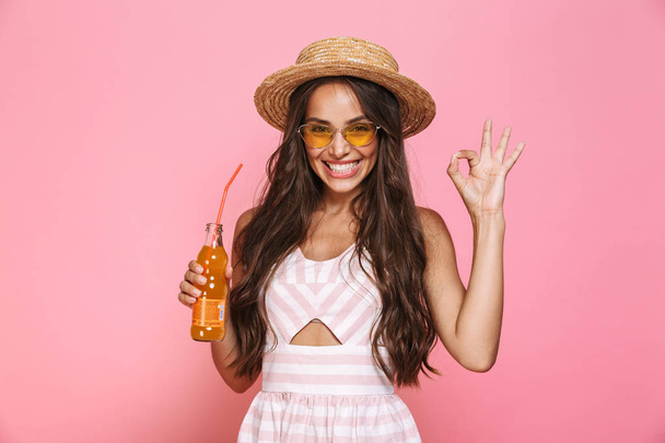 Photo of caucasian woman 20s wearing sunglasses and straw hat drinking lemonade from glass bottle isolated over pink background - Photo, image