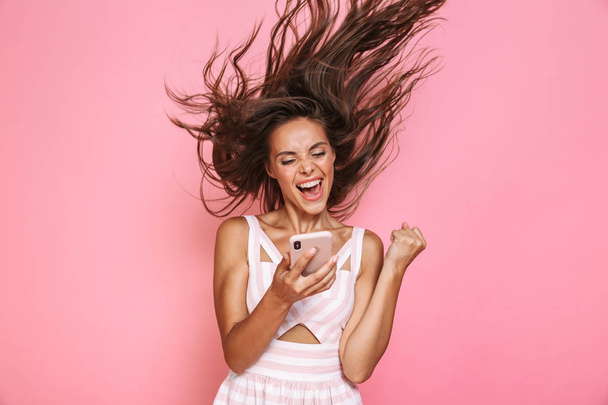 Photo of pretty woman 20s wearing dress smiling and holding smartphone with hair shaking isolated over pink background - Valokuva, kuva
