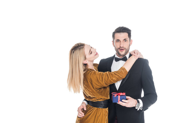 attractive woman hugging shocked boyfriend in suit with wrapped gift isolated on white - Photo, Image