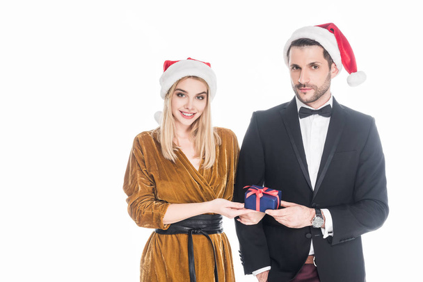 portrait of stylish young couple in santa claus hats with wrapped christmas present isolated on white - Foto, Imagen