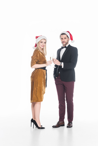side view of fashionable couple in santa claus hats clinking glasses of champagne isolated on white - Photo, Image