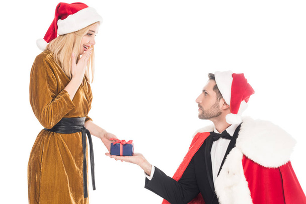 man in santa claus costume presenting gift to surprised girlfriend in santa claus hat isolated on white - Foto, immagini