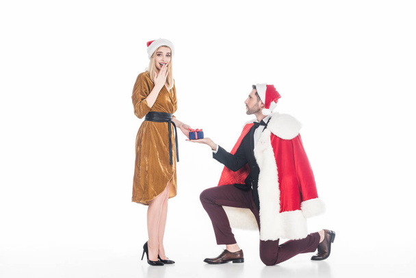 man in santa claus costume presenting gift to surprised girlfriend in santa claus hat isolated on white - Fotoğraf, Görsel