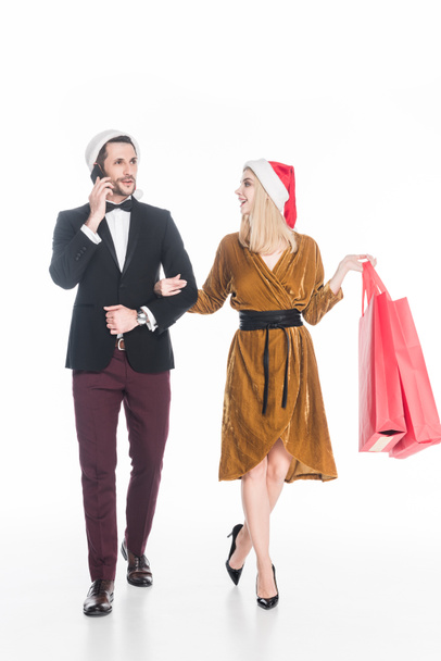 çekici bir adam akıllı kız Noel Baba şapkalı yakınındaki alışveriş torbaları üzerinde beyaz izole ile konuşurken - Fotoğraf, Görsel