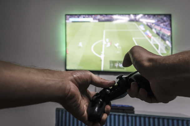 Чоловік тримає геймпад в руках перед екраном телевізора з Pro Evolution Soccer. Один гравець. Широкоекранний телевізор, що висить на стіні
 - Фото, зображення