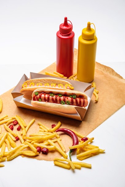 close-up shot of spicy hot dogs with french fries, mustard and ketchup on paper on white surface - Photo, Image