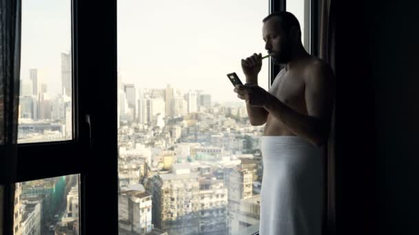 Young man in towel brushing teeth and texting on smartphone by window at home - Video