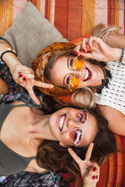 Photo from top of two joyful hippie girls smiling and lying on blanket outdoor head to head - Φωτογραφία, εικόνα