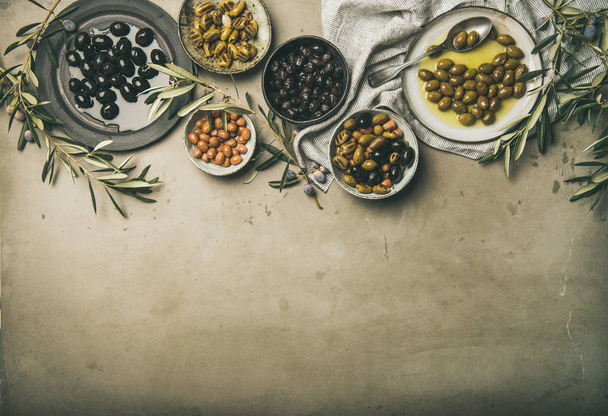 Flat-lay of various kinds of Mediterranean pickled olives in plates and bowls and olive tree branches over grey concrete table background, top view, copy space. Mediterranean meze appetizer - 写真・画像